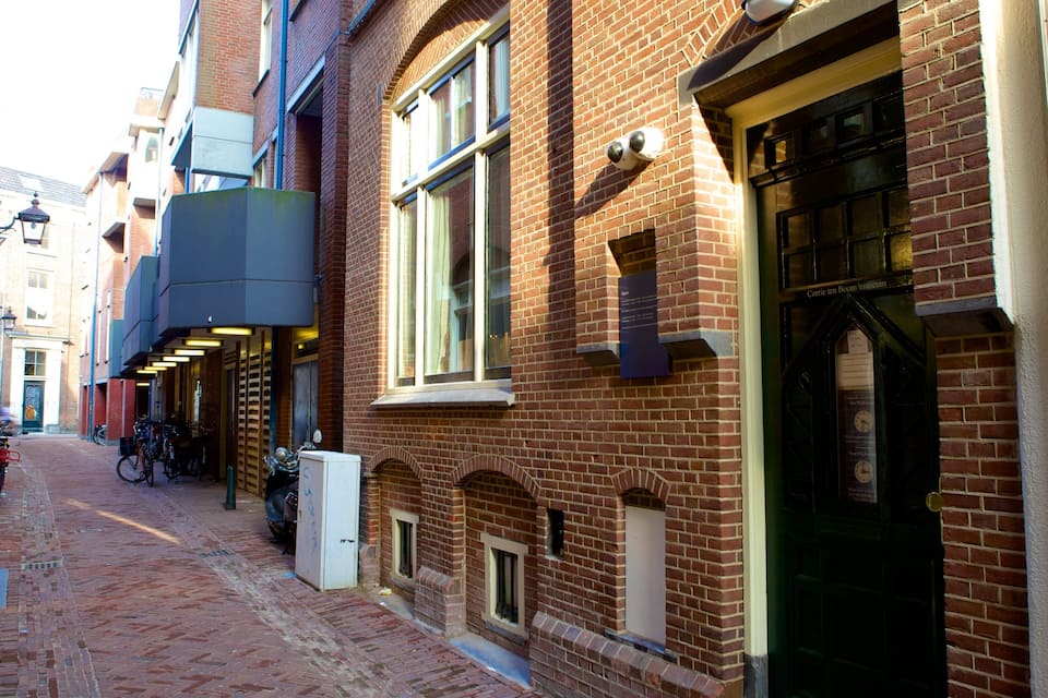 Un estrecho callejón de ladrillo con bicicletas estacionadas a un lado, un toldo de metal corrugado y un edificio con una puerta negra que dice "Corrie ten Boom Museum".