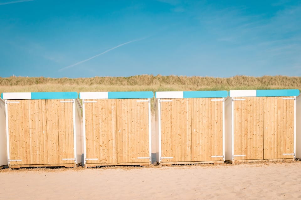 A row of wooden beach huts with blue roofs on a sandy beach.