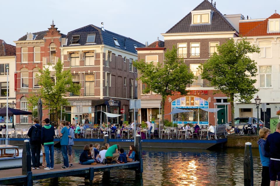 A riverside promenade with people sitting and walking, surrounded by historic buildings and a canal.