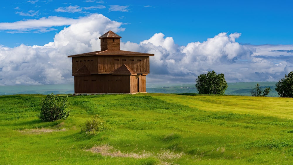 Une grange en bois, dotĂ©e dâune cheminĂ©e, se dresse sur une colline verdoyante, sous un ciel bleu parsemĂ© de nuages.