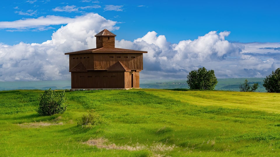 A wooden barn with a chimney on a grassy hill under a blue sky with clouds.