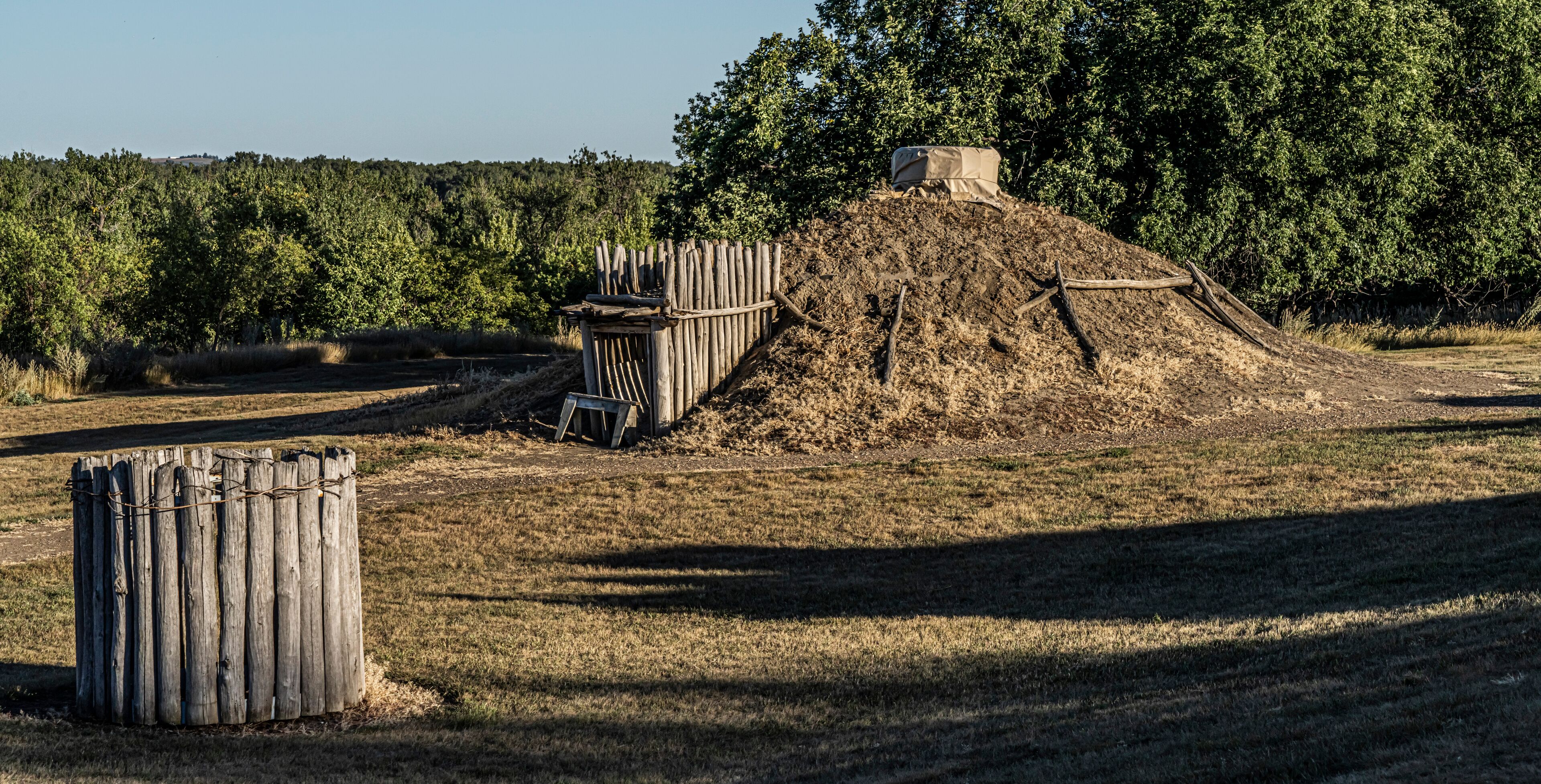 A dry, grassy field with a wooden fence, a pile of dirt, and a small structure covered with a tarp.