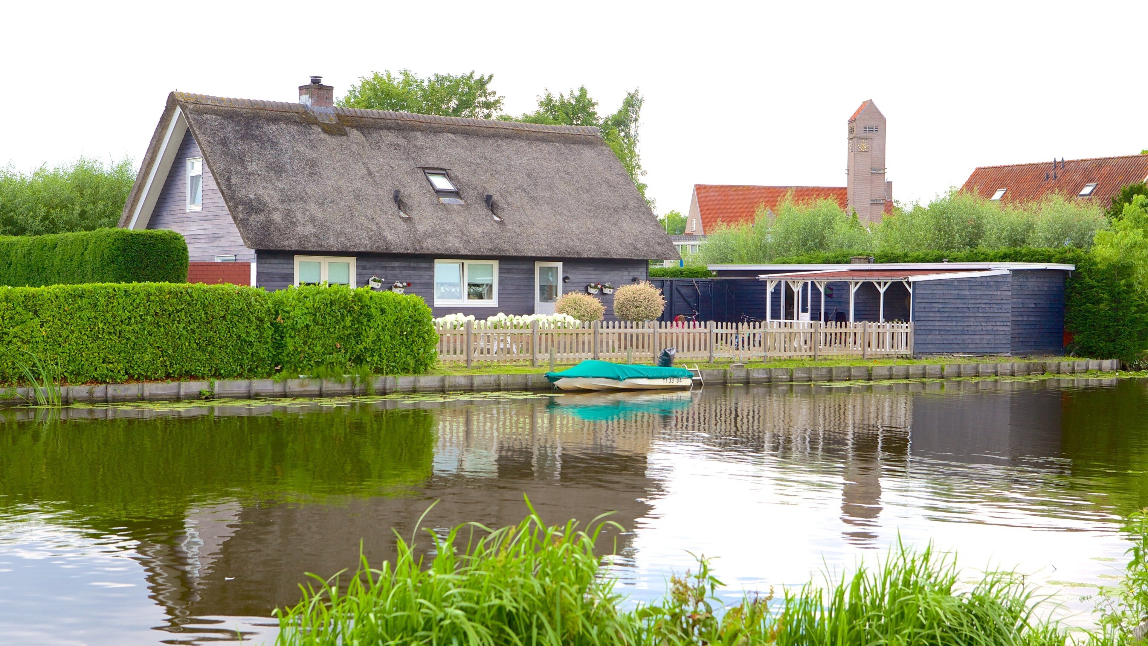 A house with a thatched roof, a small boat on a canal, and a church tower in the background.