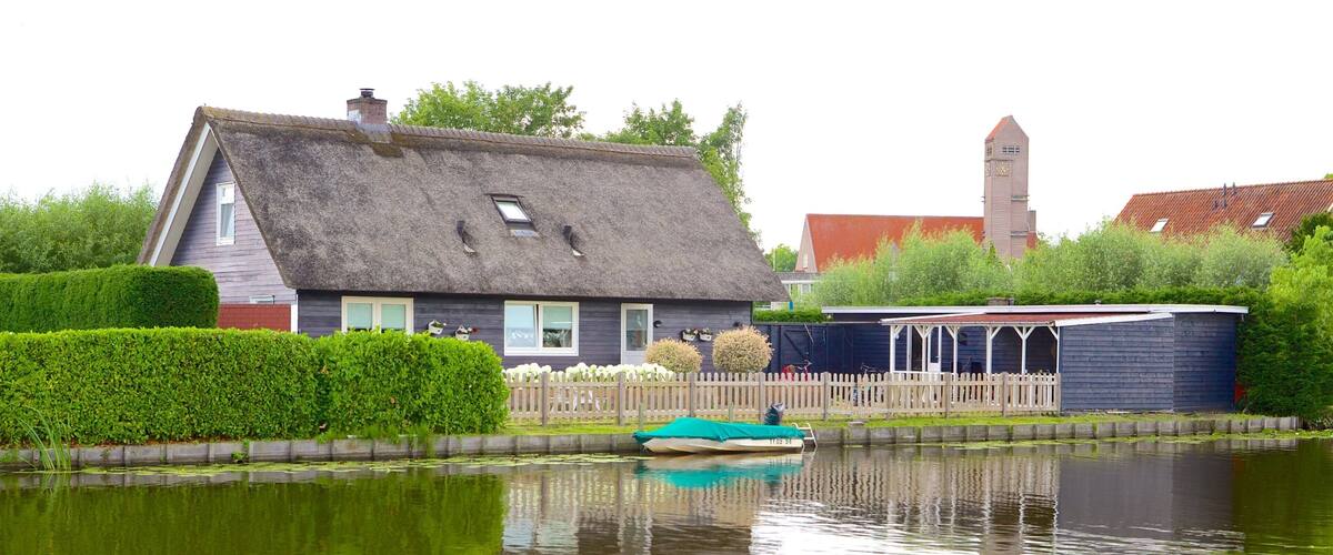 A house with a thatched roof, a small boat on a canal, and a church tower in the background.