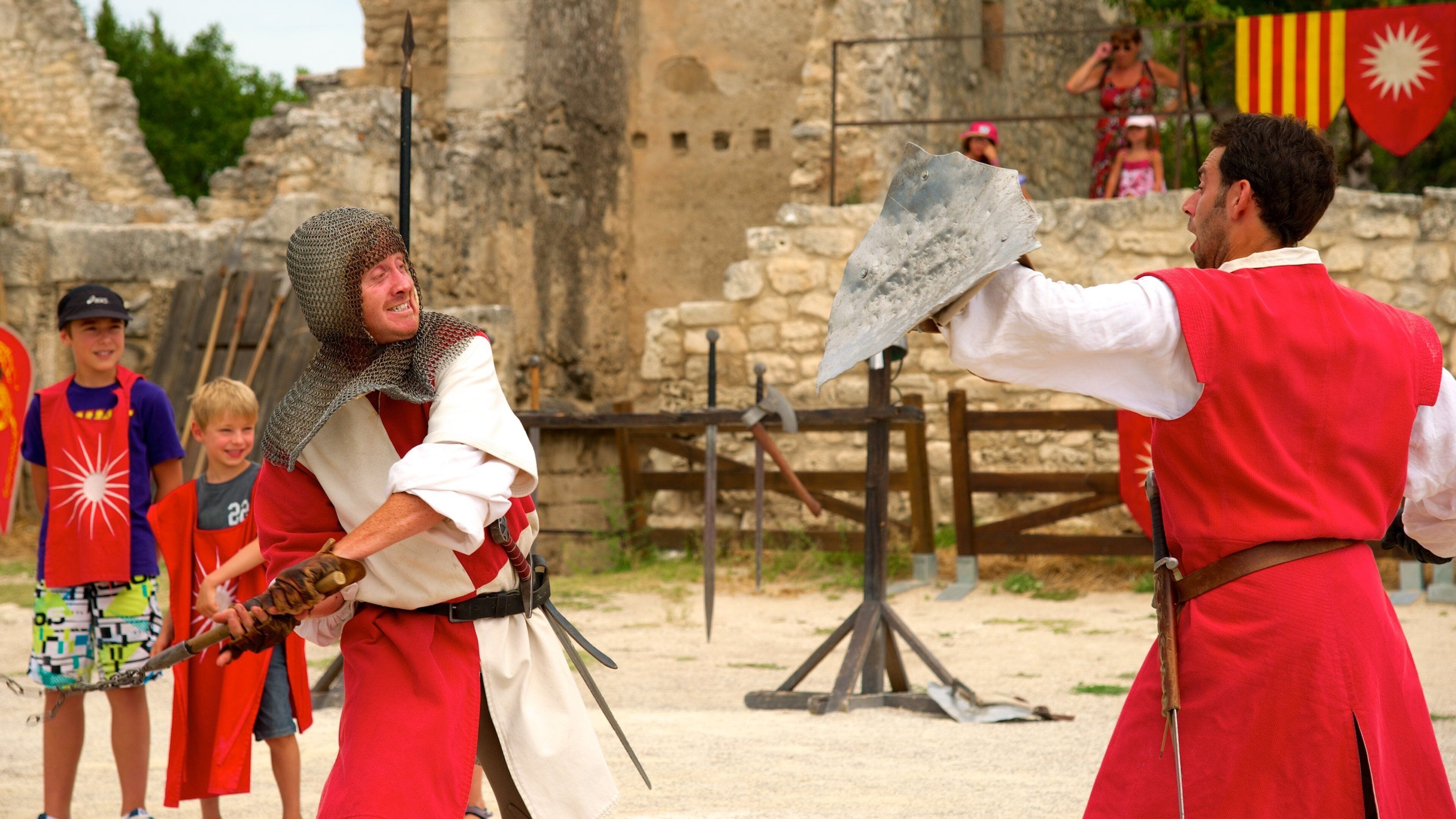 A medieval reenactment with participants in chainmail and red tunics, a stone wall, and a wooden gate in the background.