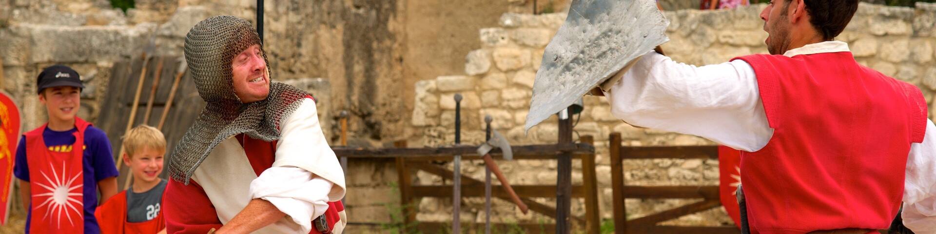 A medieval reenactment with participants in chainmail and red tunics, a stone wall, and a wooden gate in the background.