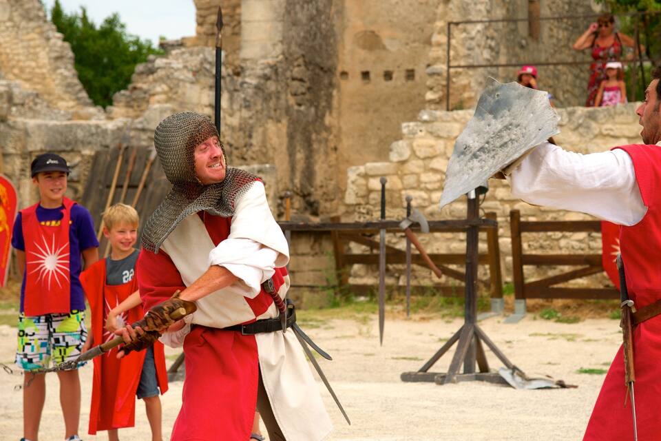 Recreación medieval con participantes con armadura de malla y túnicas rojas, un muro de piedra y una puerta de madera al fondo.