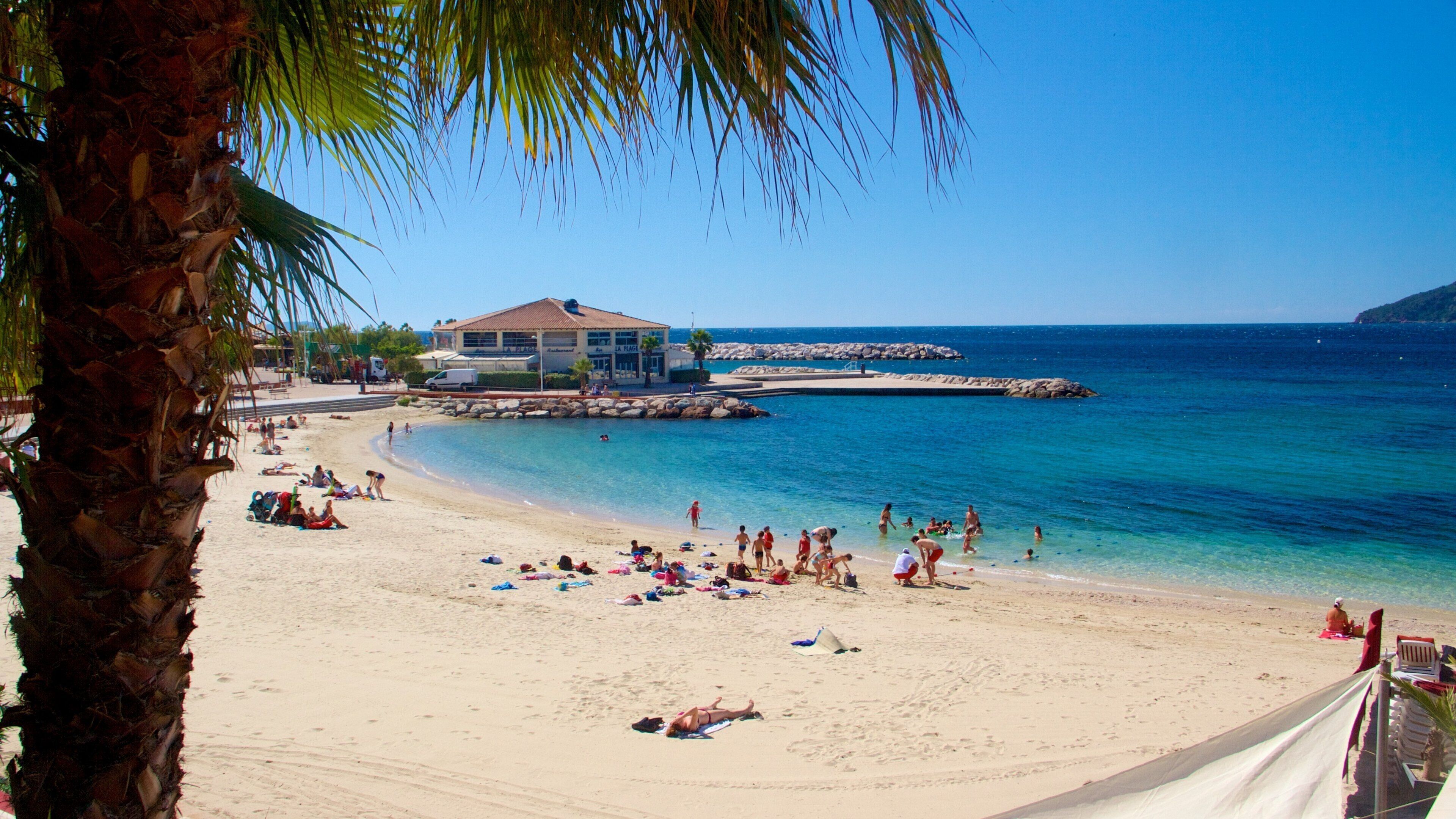 A beach with people sunbathing and swimming, a building, and rocky outcrops in the water.