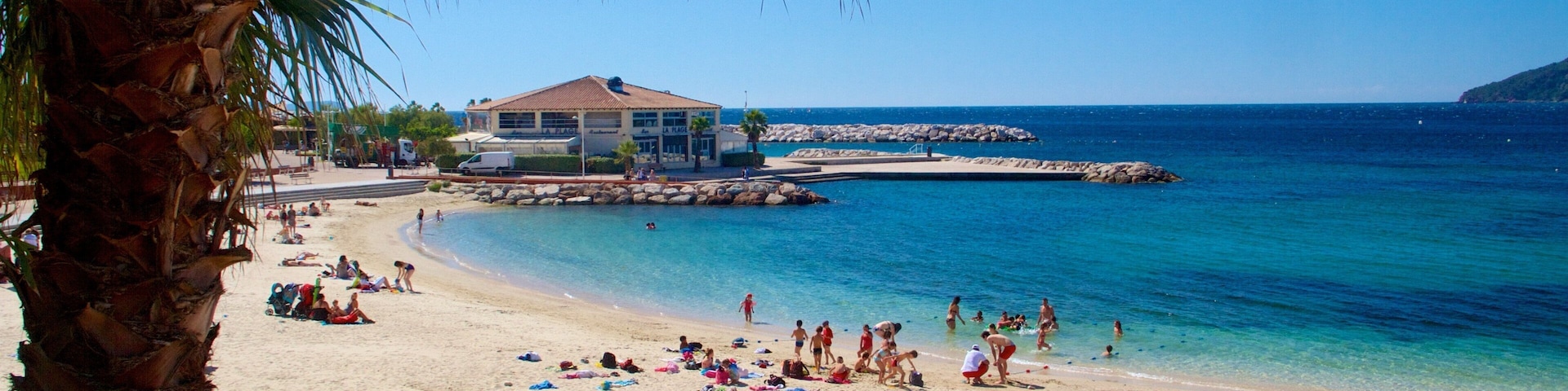 A beach with people sunbathing and swimming, a building, and rocky outcrops in the water.