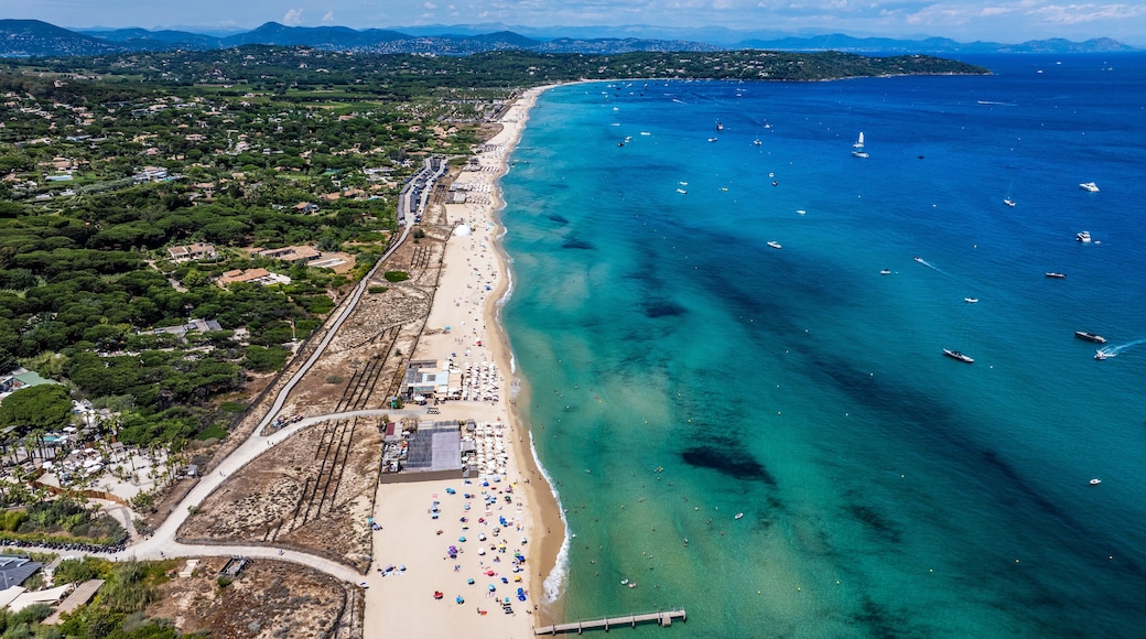 aerial view along Pampelonne Beach landscape to Saint-Tropez - located at coastline of Ramatuelle, Côte d’Azur, France - with people on the beach in water, green wooded hilly landscape in background