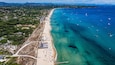 Vista aerea di una località costiera con una lunga spiaggia sabbiosa, acque blu cristalline e imbarcazioni.