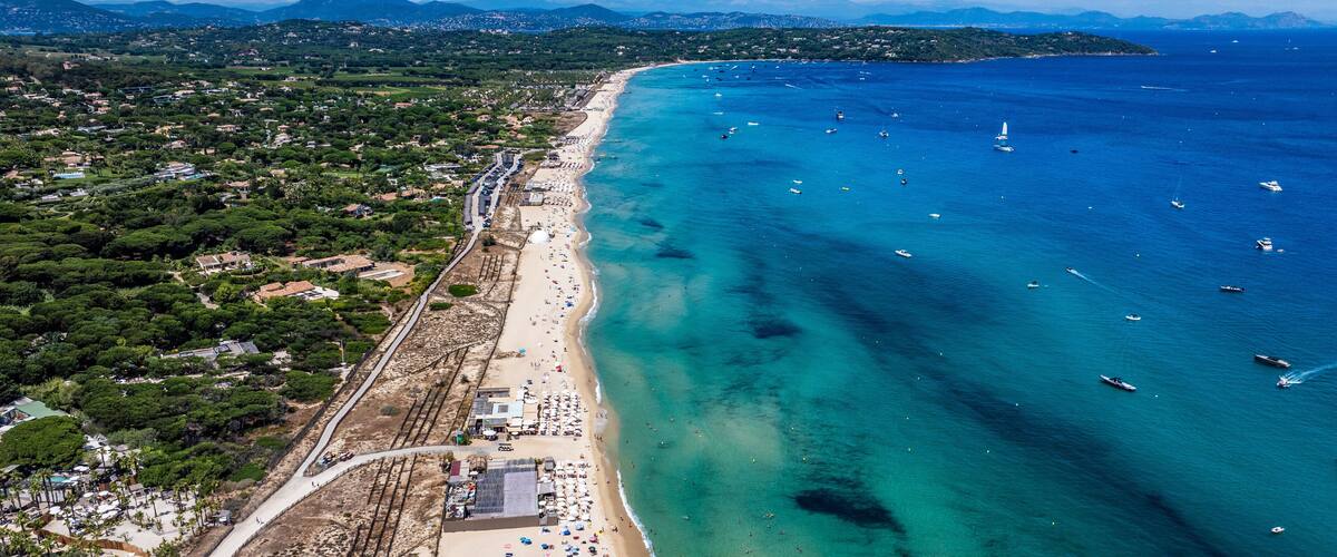 An aerial view of a coastal town with a long sandy beach, clear blue waters, and boats.