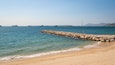 Una playa arenosa con un rompeolas rocoso y varios barcos en el agua.