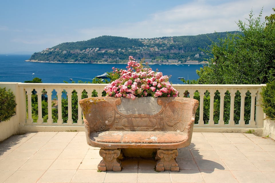 A stone bench with pink flowers in front of a coastal view.