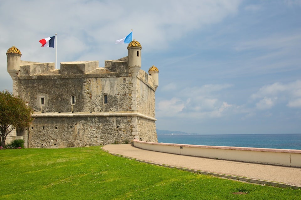 A historic stone castle with two flags, a grassy foreground, and a body of water in the background.