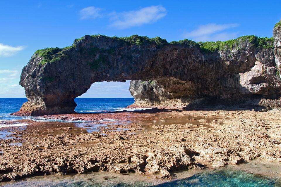 A coastal landscape with a large rock arch over the sea, a rocky beach, and clear blue waters.