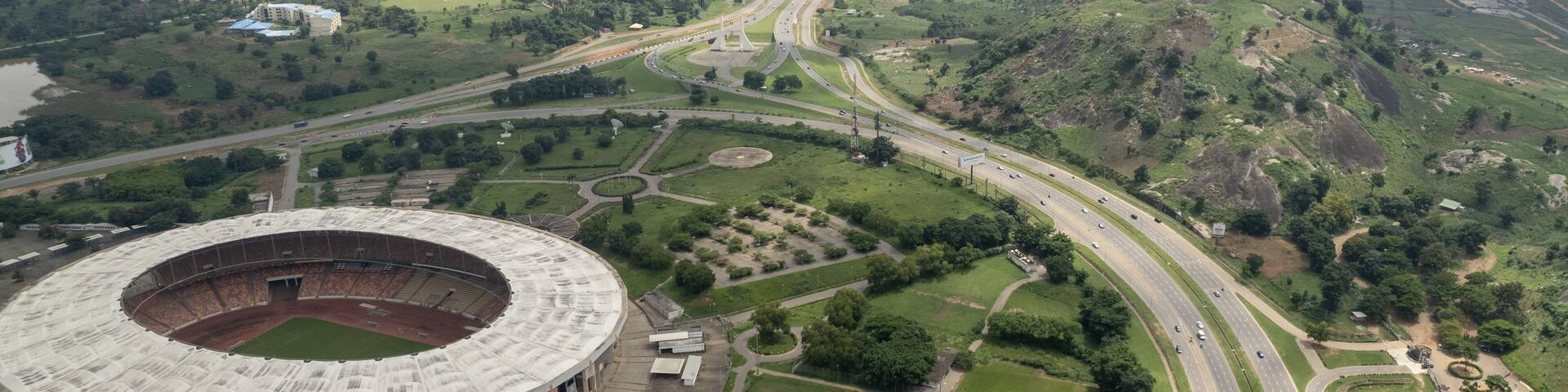 Aerial view of a large stadium nestled beside a sprawling highway, where cars stream like silver ribbons through the landscape, Abuja, Nigeria.