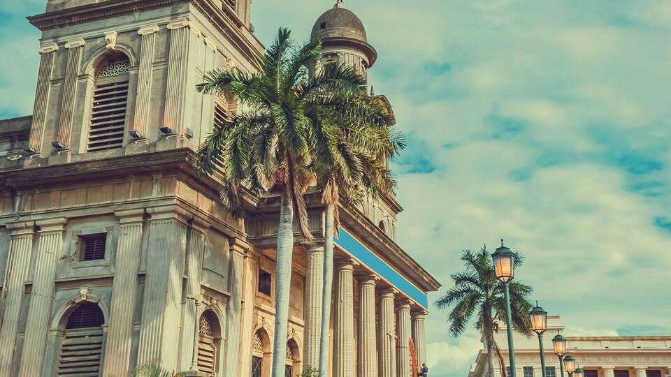 A historic building with a dome and bell tower, palm trees, and a paved walkway.