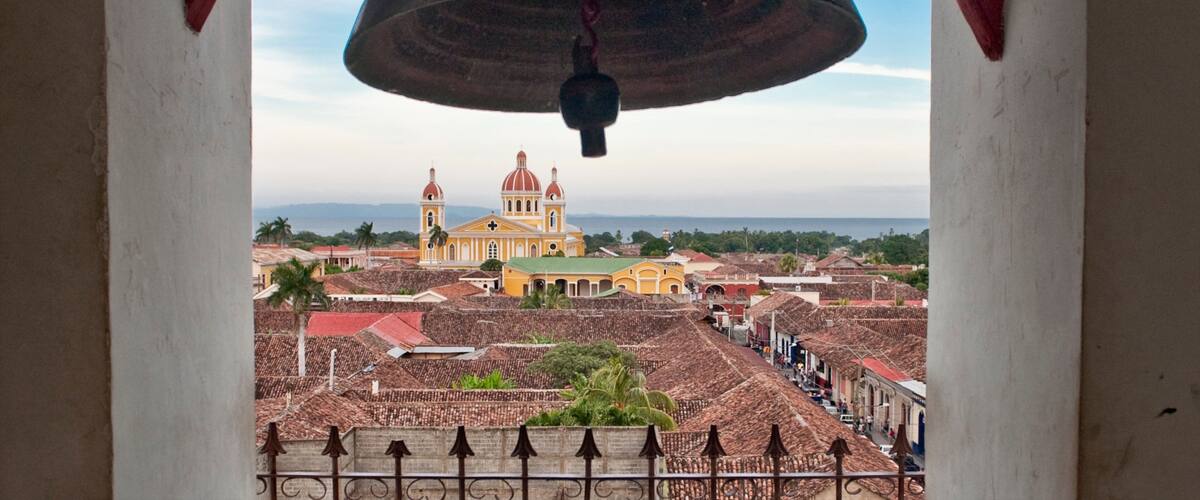 A large bell hanging from a structure, overlooking a town with colonial architecture and a prominent church with red domes.