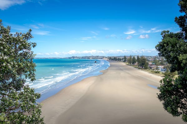 Uma extensa praia de areia com águas azul-claras, cercada por vegetação e edifícios ao longe.