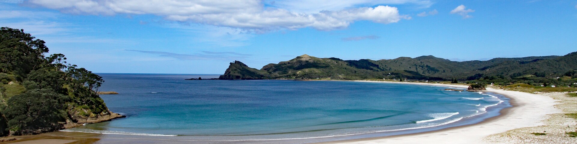 A coastal landscape with a sandy beach, clear blue waters, and green hills in the background.