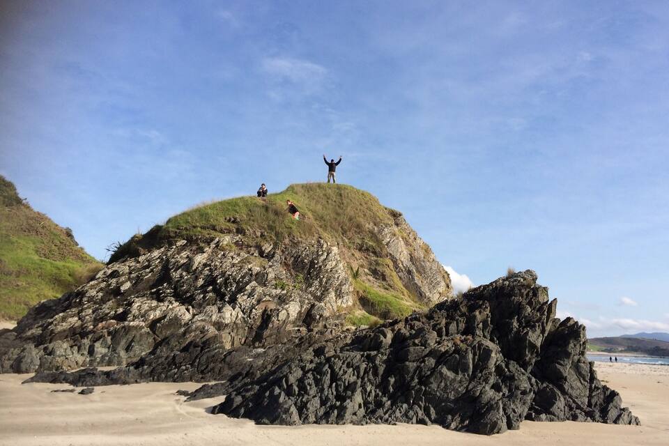 Een rotsachtige heuvel met mensen erop, een strand op de voorgrond en een heldere blauwe lucht.