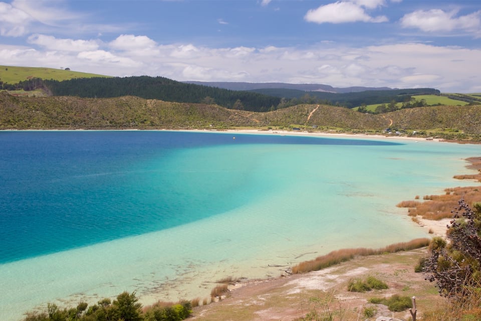 A coastal landscape with clear turquoise waters, sandy beaches, and rolling hills covered in greenery.