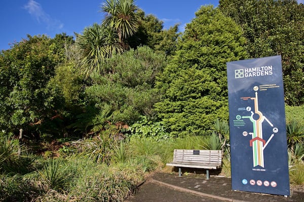 A sign for Hamilton Gardens with a map and a bench in the foreground.