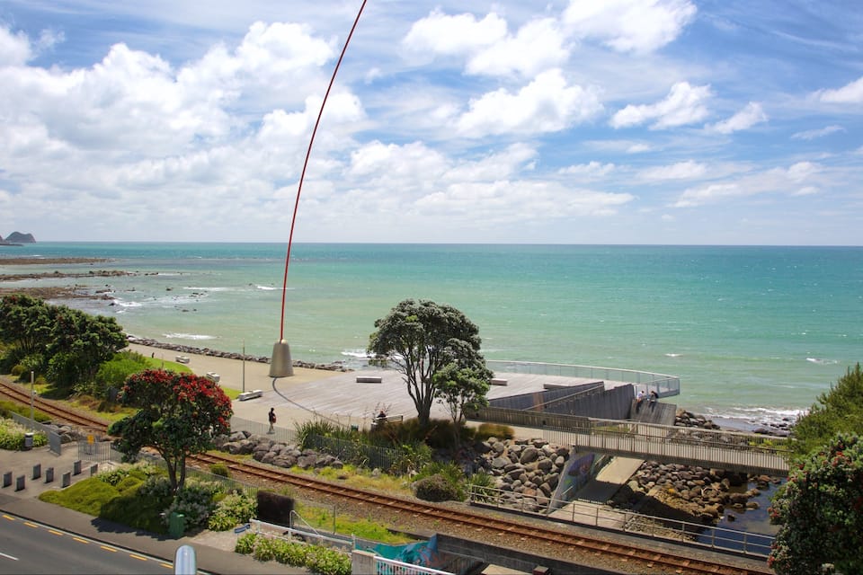 Paisagem costeira com praia, trecho rochoso, uma árvore e uma linha de trem.
