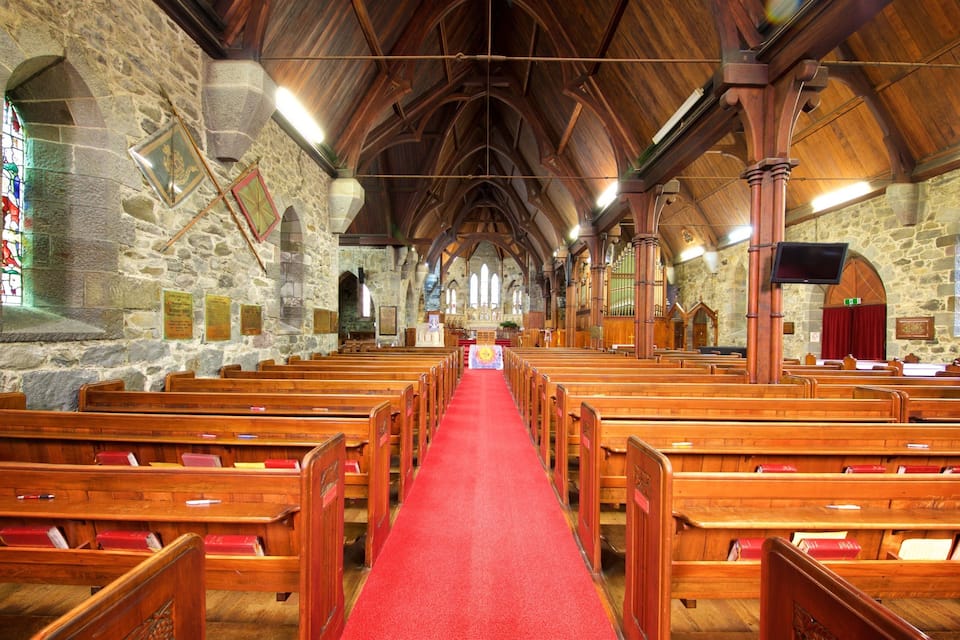 A church interior with wooden pews, a red carpet aisle, stone walls, and a wooden ceiling.