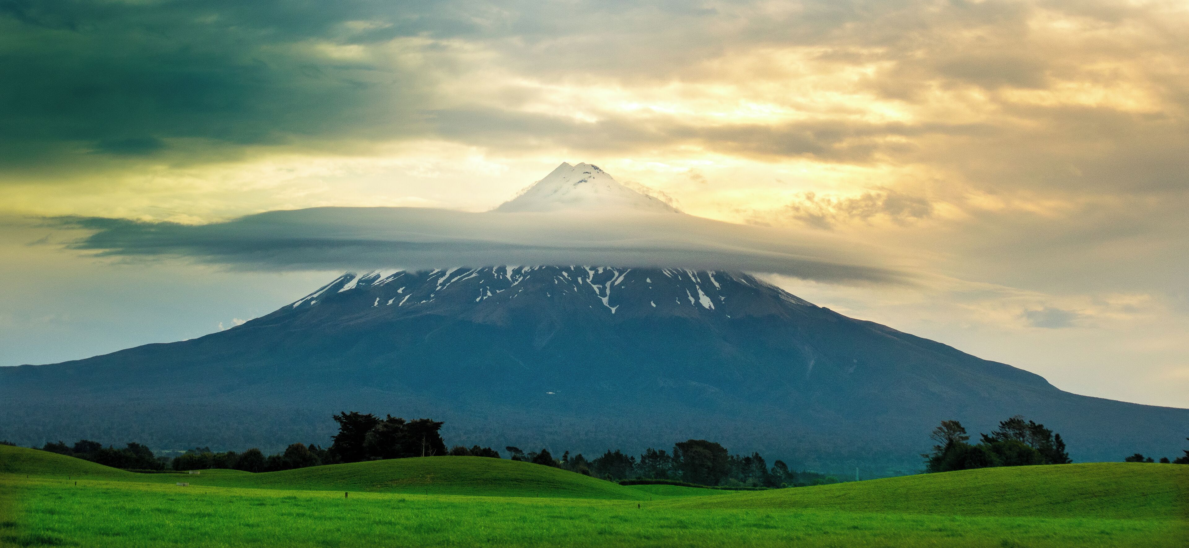 A snow-capped mountain with a grassy foreground and a cloudy sky.