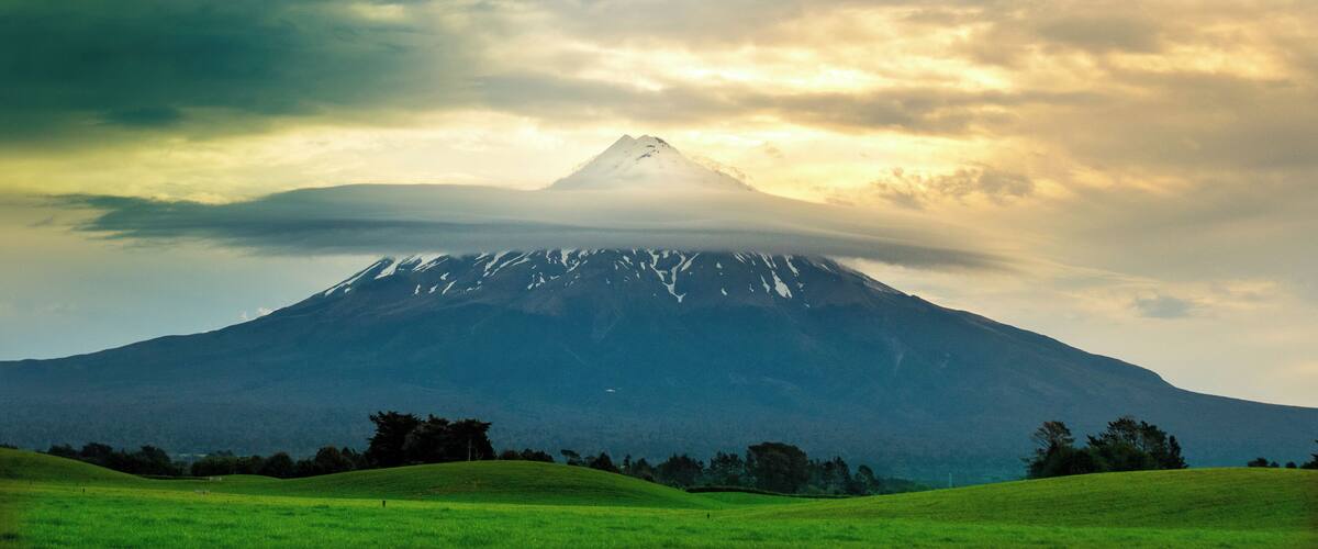 A snow-capped mountain with a grassy foreground and a cloudy sky.
