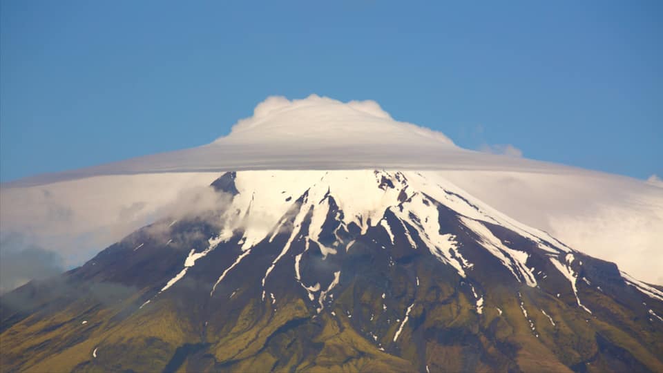 A snow-capped mountain with a cloud base, surrounded by green valleys.