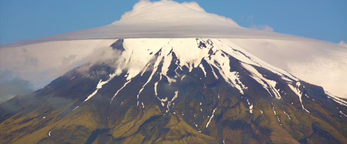 A snow-capped mountain with a cloud base, surrounded by green valleys.