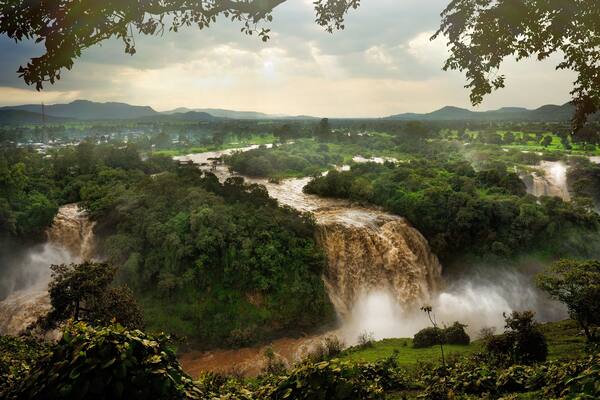 A waterfall cascading through a lush valley surrounded by dense forest and distant hills.