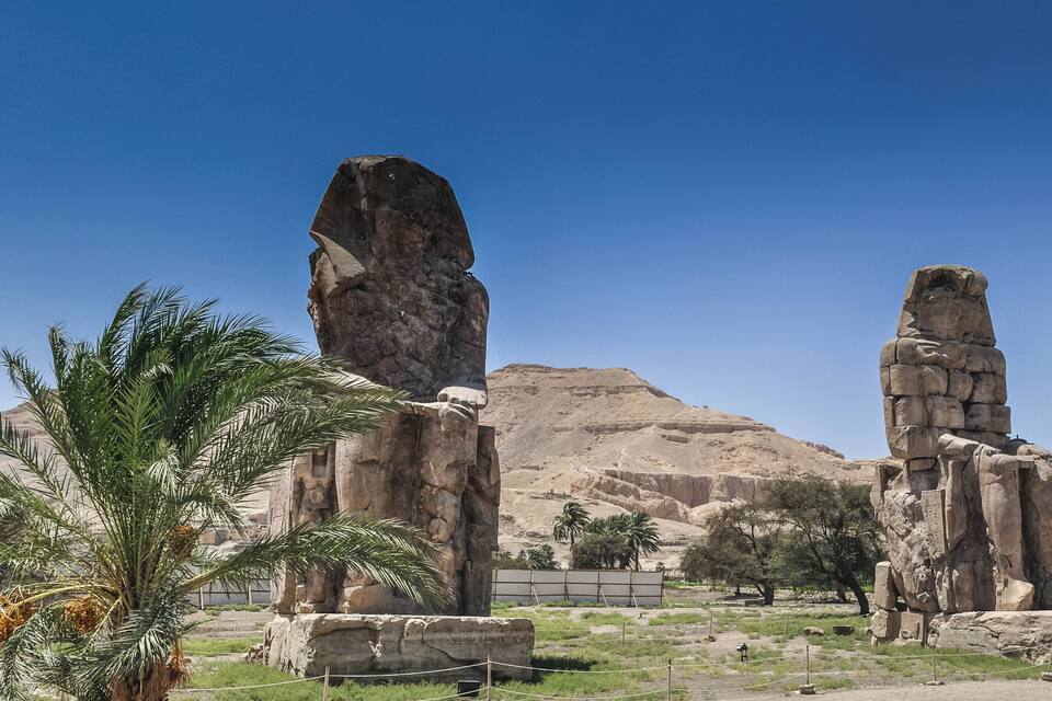 A desert landscape with two large stone statues and palm trees.