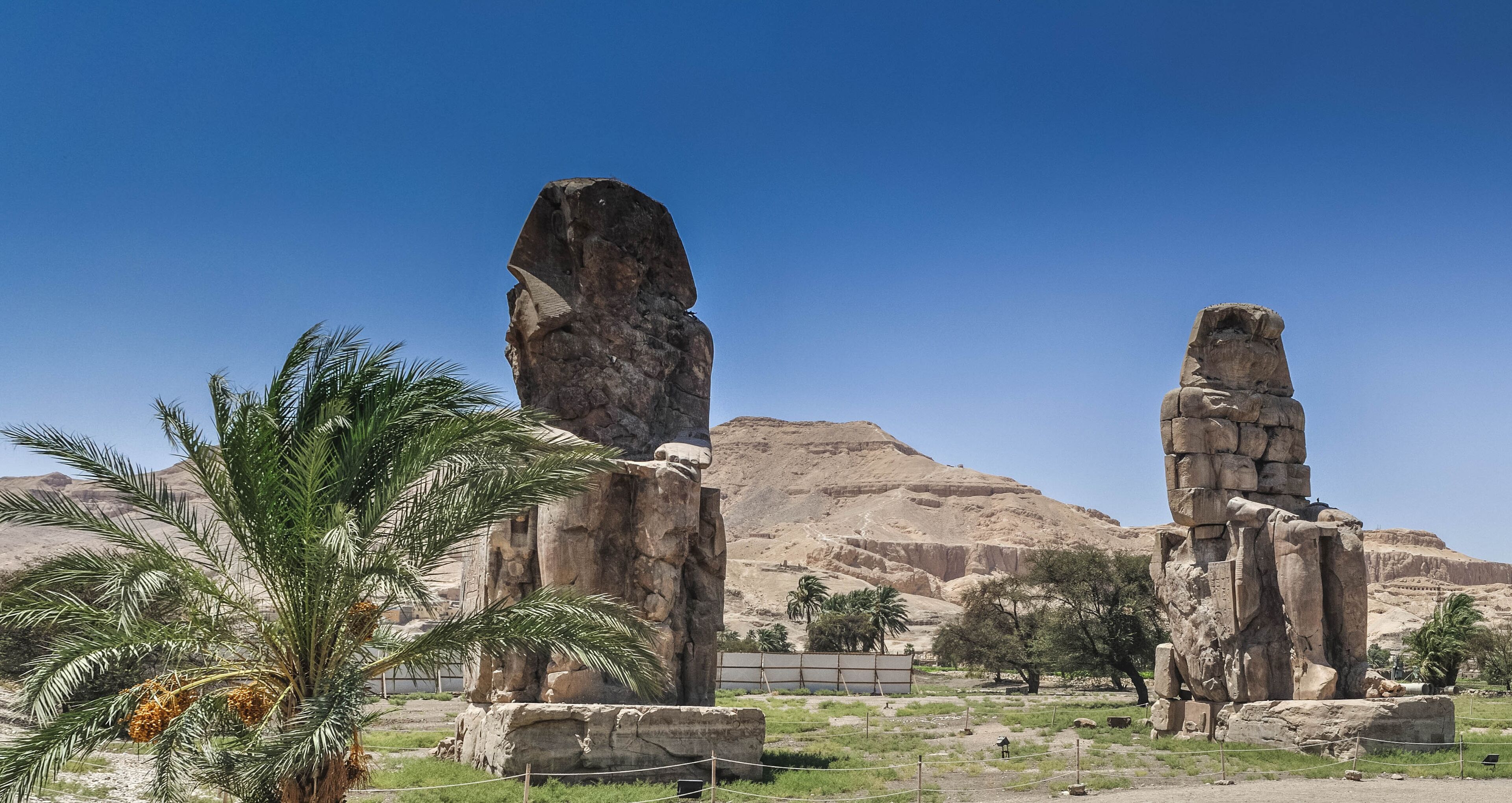 A desert landscape with two large stone statues and palm trees.