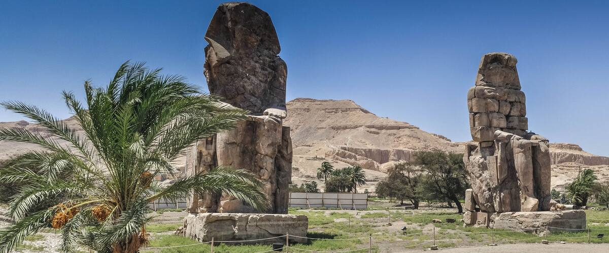 A desert landscape with two large stone statues and palm trees.