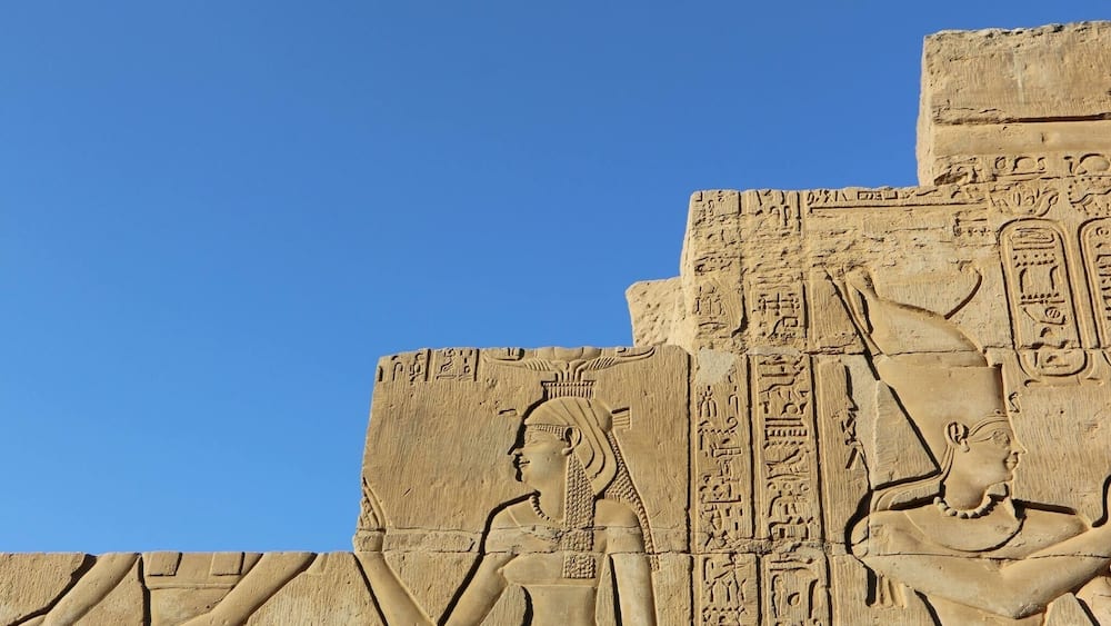 A large stone relief with hieroglyphics and two figures, one seated and one standing, against a clear blue sky.