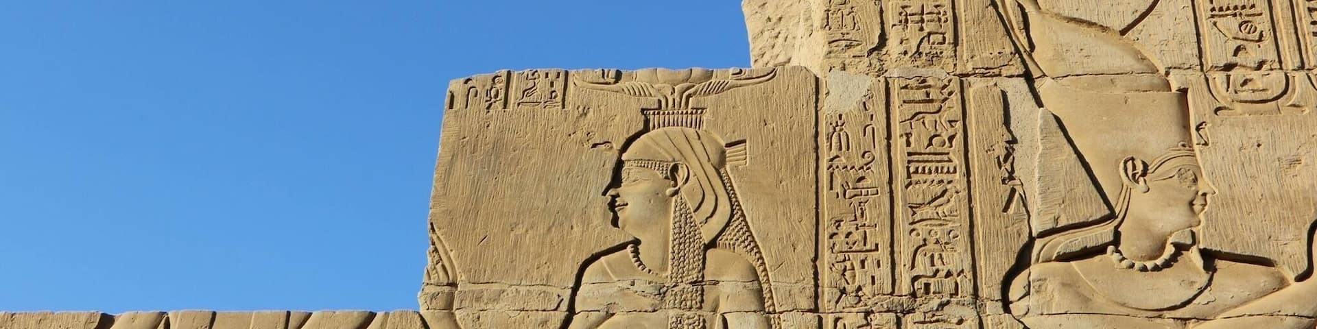 A large stone relief with hieroglyphics and two figures, one seated and one standing, against a clear blue sky.