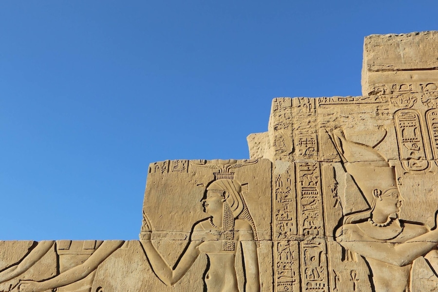 A large stone relief with hieroglyphics and two figures, one seated and one standing, against a clear blue sky.