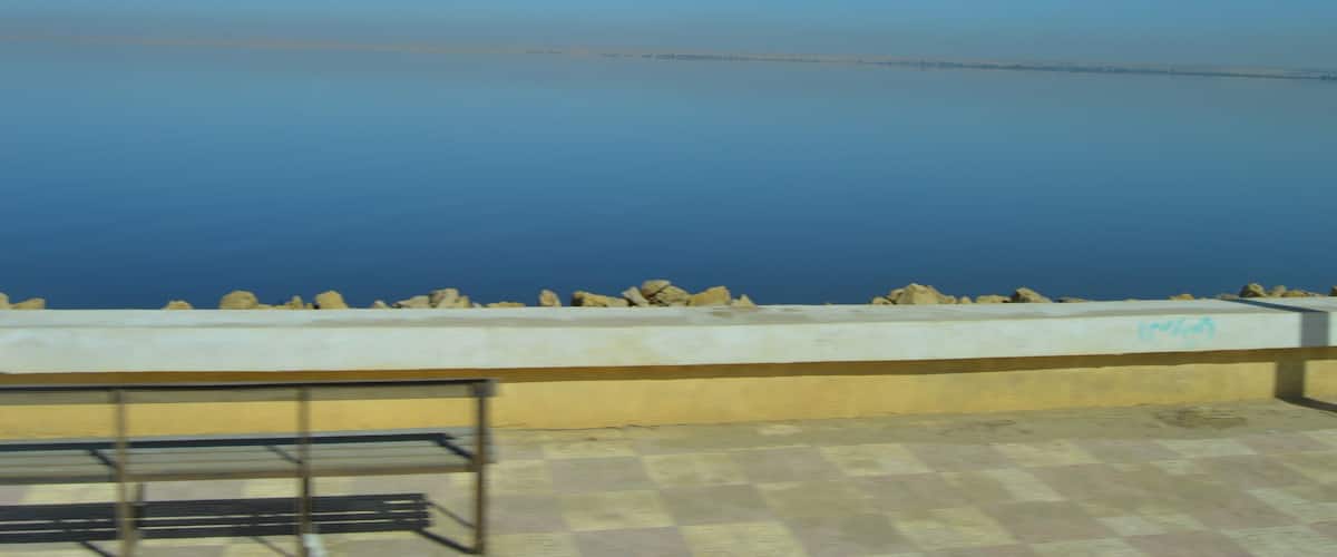 A calm body of water with a distant shoreline, a concrete barrier, and a metal railing.