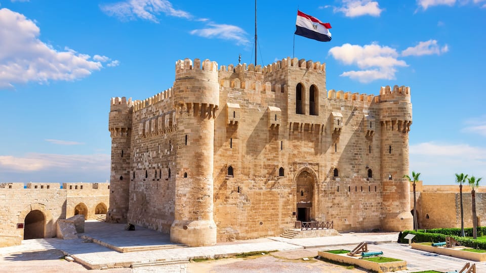 Um castelo de pedra histórico com uma bandeira, portões em arco e um pátio com canhões.