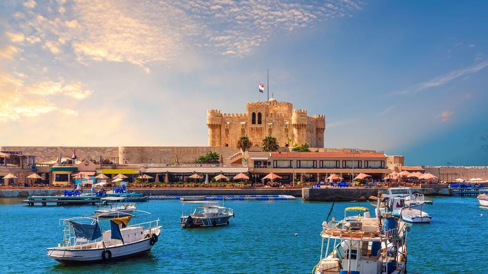 Um castelo histórico com uma bandeira, um porto com barcos e um calçadão à beira-mar.