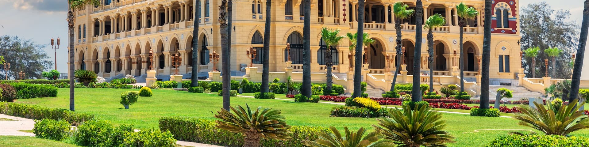 A grand, ornate building with multiple towers and arches, surrounded by well-maintained gardens and palm trees.