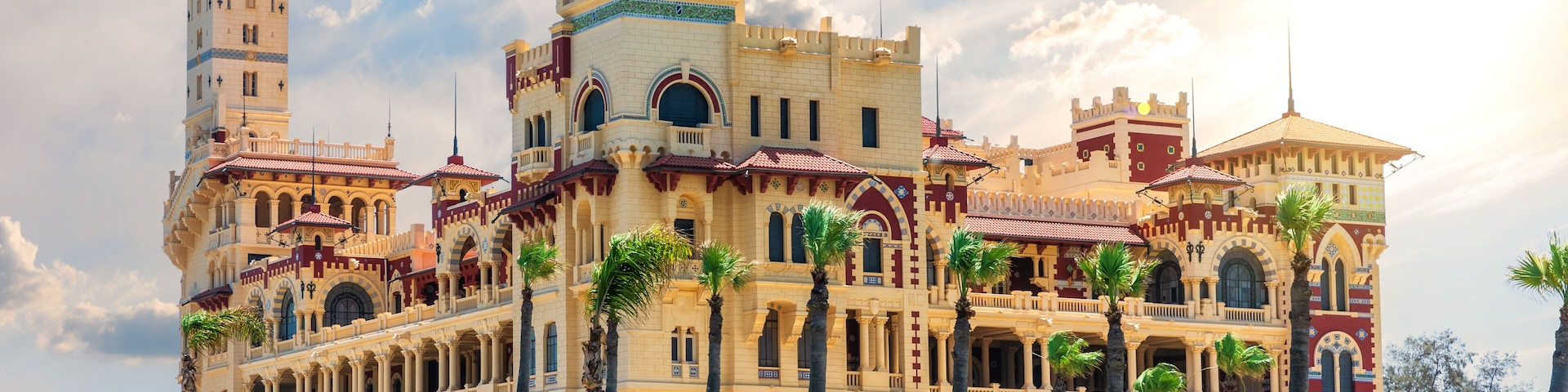 Un edificio imponente y ornamentado con múltiples torres y arcos, rodeado de jardines cuidados y palmeras.