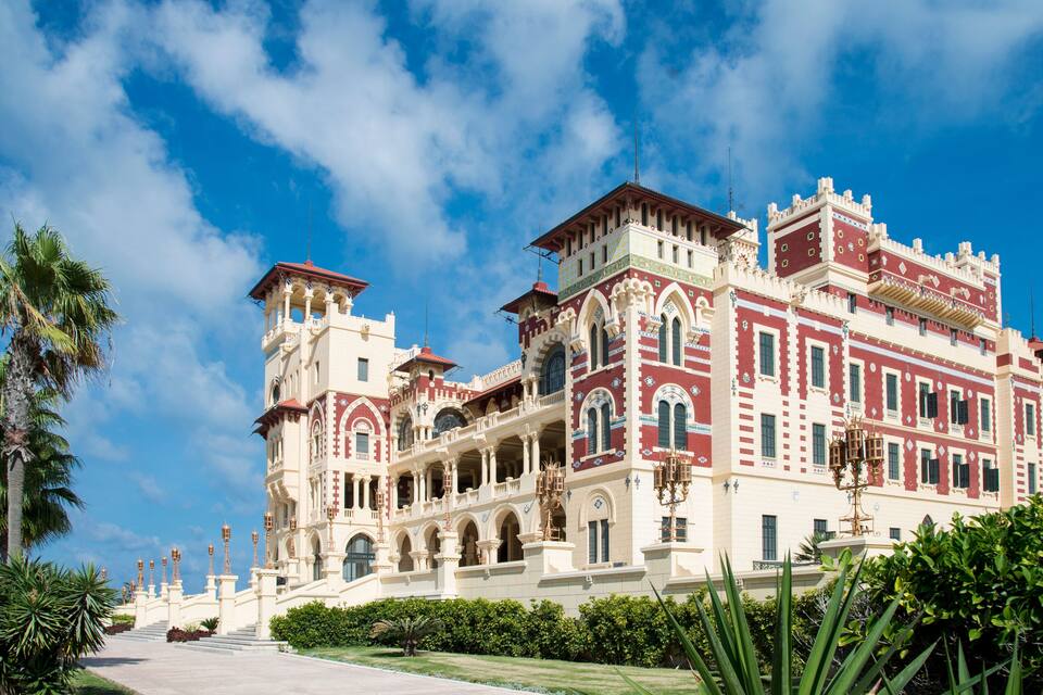 Un edificio majestuoso y ornamentado con múltiples torres y balcones, rodeado de abundante vegetación y palmeras.