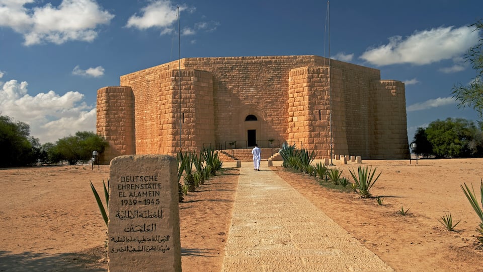 A historical building with a central archway, surrounded by a paved walkway and small plants.