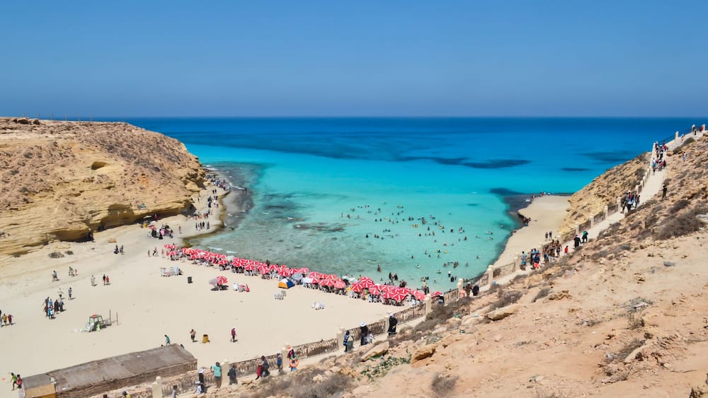 A coastal beach with clear turquoise waters, a sandy shore, and rocky cliffs.