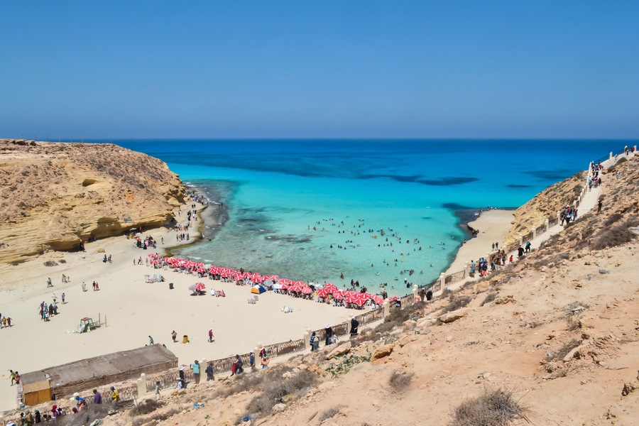 Una playa costera con aguas turquesas cristalinas, una orilla arenosa y acantilados rocosos.